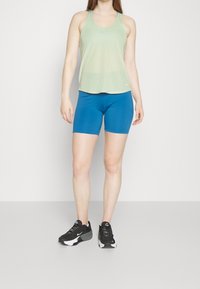 Ljusgrön nät-tanktop med rund halsringning, kombinerad med åtsittande blå shorts. Svarta träningsskor med vita accenter. Släta texturer.