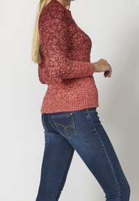 Knitted sweater in rose gradient, textured pattern, round neckline. Paired with blue denim jeans featuring decorative stitching on pockets.