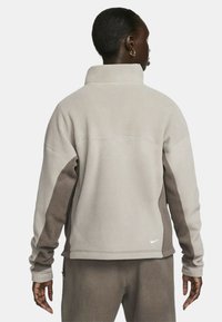 Giacca in pile Nike in tonalità di grigio chiaro e marrone, caratterizzata da un colletto alto, maniche lunghe e un logo discreto sulla parte inferiore della schiena. Texture morbida.