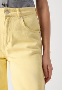 Gros plan d'une personne portant un jean taille haute jaune pâle avec des détails de boutons et de poches, et un haut gris rentré à l'intérieur.