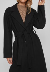 Γυναίκα που φοράει ένα μαύρο παλτό με ζώνη πάνω από μια υφασμάτινη μπεζ μπλούζα με ψηλό λαιμό, σηκώνοντας το δεξί της χέρι με τα δάχτυλα ελαφρώς καμπυλωμένα.