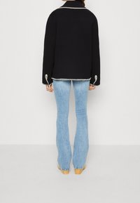 Zwarte jas met witte stiksels, brede lichtblauwe flare jeans en houten klompen, bekeken vanaf de achterkant.