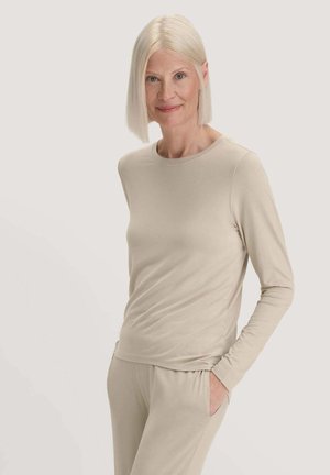 Reife Frau mit kurzen blonden Haaren, die ein beiges Langarmoberteil und passende Hosen trägt, stehend mit einer Hand in der Hosentasche vor einfarbigem Hintergrund.