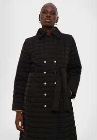 Melnā iešūta garā mēteli ar jostu, izteiktiem pogu akcentiem un apkakli. Tam ir teksturēts audums un sānu kabatas.