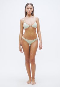 Σετ μαγιό σε ανοιχτό πράσινο χρώμα με τοπ σε σχήμα τριγώνου και μαγιό κάτω μέρος με χαμηλή μέση. Ομαλή υφή με ρυθμιζόμενους ιμάντες και ελάχιστη κάλυψη.