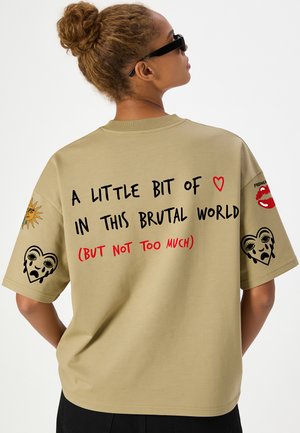 Frau mit Sonnenbrille und Dutt trägt ein beiges Hemd mit der Aufschrift "A little bit of love in this brutal world (but not too much)" mit Herz und künstlerischen Designs.