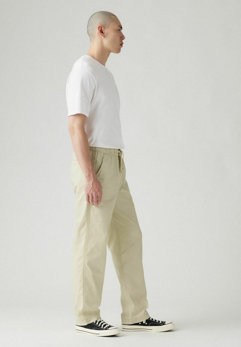 Pantalones beige con cordón y bolsillos laterales, combinados con una camiseta blanca de manga corta y zapatillas negras de caña baja. Tela suave, corte relajado.