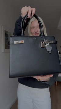 Fekete bőr kézitáska dupla fogantyúval, aranyszínű fém kiegészítőkkel és zárható zárral. Sima felület és strukturált forma; levehető kulcstartóval.