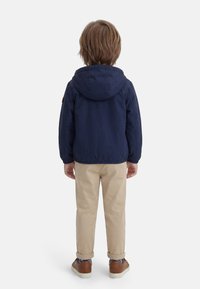 Παιδί που στέκεται με την πλάτη, φοράει μπλε μαρινό φουτάκι με κουκούλα, μπεζ παντελόνια με μανσέτες, μπλε κάλτσες με σχέδιο και καφέ παπούτσια σε λευκό φόντο.