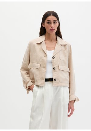Woman wearing beige buttoned jacket over white top and high-waisted white pants, standing with one hand in pocket against plain background.
