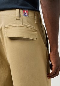 Khaki bukser med en lomme bagpå, prydet med et Wrangler-mærke. Stoffet er glat med en let glans og har en skræddersyet pasform.