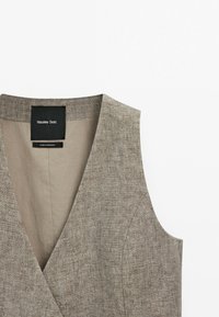 Beżowa kamizelka lniana z dekoltem w kształcie litery V, bez rękawów, o subtelnej fakturze i jasnym beżowym podszewczem. Widoczna metka Massimo Dutti przy dekolcie.