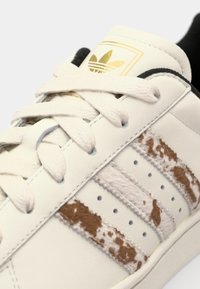 White leather sneaker featuring brown and cream patterned accents, a textured finish on the stripes, and a gold logo on the tongue.