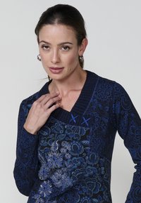 Stricktop in einem tiefen Marineblau, mit floralen Mustern in verschiedenen Blautönen, einem strukturierten Kragen und dekorativen Stickereien.