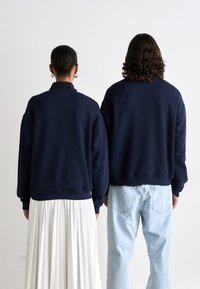 Zwei Personen stehen Rücken an Rücken, tragen marineblaue Sweatshirts; eine in einem weißen plissierten Rock, die andere in hellblauen Jeans, vor einem schlichten Hintergrund.