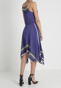 Žena v bezrukávových modrých šatech midi s potištěným vzorem a spodním lemem ve tvaru kapesníku, stojící bokem. Také má na sobě černé sandály s blokovým podpatkem a kotníkovým páskem.