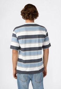 Persoon met krullend haar, die een kortemouwshirt draagt met blauwe, witte en grijze strepen, en lichte spijkerbroek, met het gezicht naar weg gekeerd tegen een effen achtergrond.