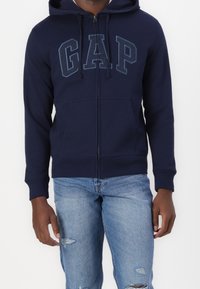 Sweat à capuche zippé bleu marine avec cordon de serrage, poches avant, et logo GAP en bleu clair contrastant. Associé à un jean bleu clair déchiré.