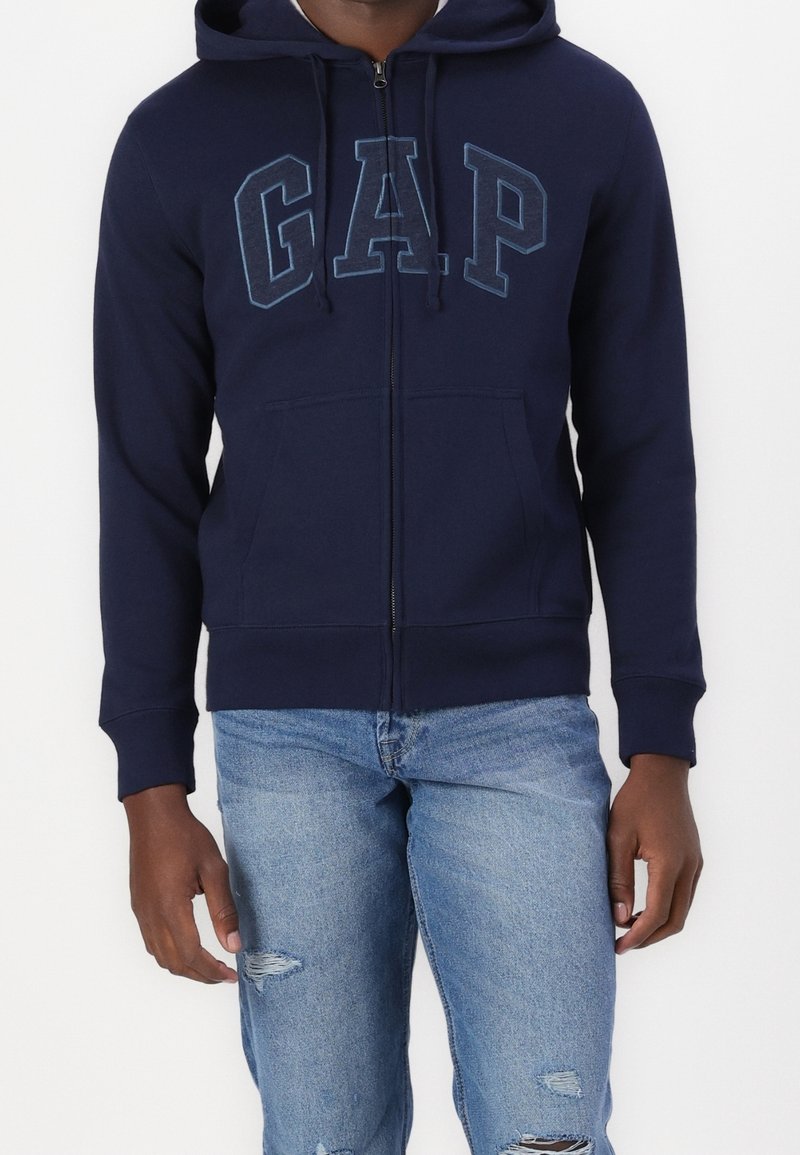 Sweat à capuche zippé bleu marine avec cordon de serrage, poches avant, et logo GAP en bleu clair contrastant. Associé à un jean bleu clair déchiré.