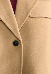 Beige wollen blendjas met een klassieke notch-lapel, sluiting met één zwarte knoop en een zachte textuur met een gestructureerde silhouet.