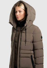 Pufferjack in taupe met een grote capuchon, voorzien van een ritsluiting, zwarte accenten en een geborduurd patroon. Zijzakken met ritsen.