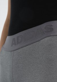 Detailní záběr na osobu, která má na sobě šedé legíny Adidas s širokým elastickým pasem a černý přiléhavý top.