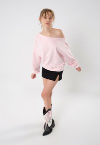 Sweatshirt oversize rosa chiaro con design off-the-shoulder, abbinato a una minigonna nera con spacco laterale. Stivali cowboy decorativi bianchi e neri.