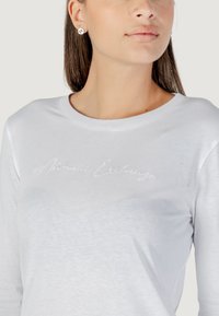 T-shirt bianco a maniche lunghe con tessuto leggermente testurizzato e logo ricamato "Armani Exchange" in stile corsivo.