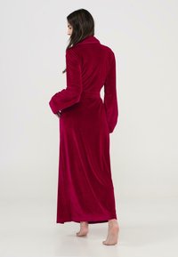 Bata de terciopelo a la altura de la rodilla en un rico burdeos con cuello chal y cintura ceñida. Presenta mangas largas y un diseño fluido y drapeado.