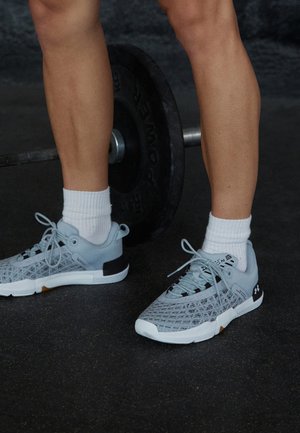 Beine mit weißen Socken und grauen Sportschuhen stehen auf dem Hallenboden in der Nähe einer Langhantel mit schwarzen Gewichtsscheiben.