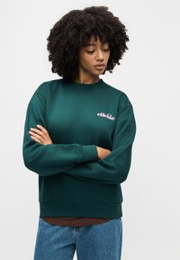 Γυναίκα με σγουρά μαλλιά που φοράει σκούρο πράσινο φούτερ με το λογότυπο "ellesse", μπλε τζιν, έχει σταυρωμένα τα χέρια και κοιτάζει προς τα κάτω.