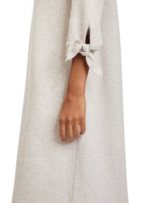 Bras de personne portant une robe beige texturée à manches trois-quarts, nouée en un nœud au poignet, main détendue et doigts légèrement courbés.