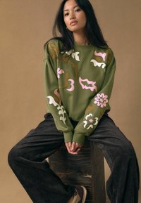 Sweat-shirt vert oversize avec des détails de broderies florales et abstraites colorées, associé à un pantalon en velours côtelé noir et des chaussures beige.