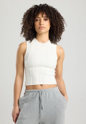 Weißes, geripptes ärmelloses Crop-Top mit hohem Kragen und strukturierten Streifen, kombiniert mit grauen Jogginghosen mit Kordelzug.