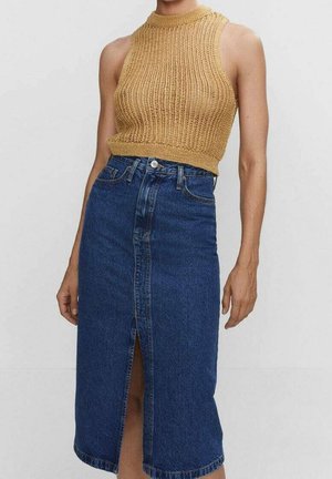 Femme portant un haut court en tricot sans manches couleur beige et une jupe midi en jean bleu taille haute avec une fente sur le devant.