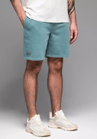 Shorts de sport bleu clair avec poches latérales, texture lisse et petite étiquette de marque sur la jambe gauche, associés à des baskets beiges.