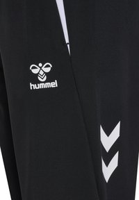 Primo piano di pantaloni sportivi neri con logo Hummel ape bianco e due strisce bianche a forma di chevron sulla gamba.