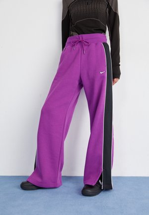 Persoon draagt losse paarse broek met zwarte zijstrepen, een zwarte gestructureerde lange mouwen top en zwarte schoenen, staand op een blauwe vloer.