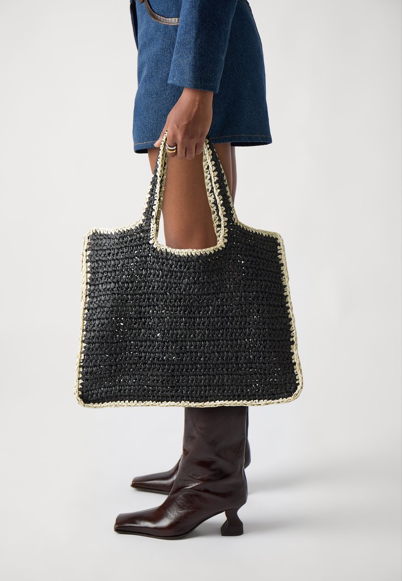 Personne vêtue d’une tenue bleue tenant un grand sac cabas noir tissé avec des bords crème, portant des bottes à talons marron foncé, sur un fond uni.
