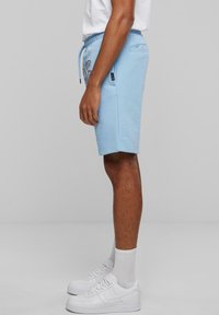 Pantalones cortos de color azul claro, fabricados en una tela suave, con cintura ajustable mediante cordón, bolsillos laterales y un diseño gráfico audaz. Combinados con zapatillas blancas y calcetines.