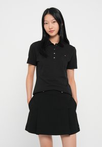 Sort polo-shirt med krave og fem knapper; parret med en sort plisseret nederdel med guld accenter og lommer.