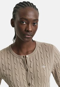 Beige Kabelstrick-Cardigan mit rundem Ausschnitt, Knopfleiste und gesticktem Logo. Weiche Textur mit erhabenen Musterdetails.