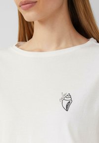 Weißes Baumwoll-T-Shirt mit Rundhalsausschnitt, das auf der linken Seite ein kleines schwarzes Muscheldesign und den Text "cabin" aufgedruckt hat.