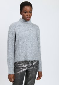 Kvinde iført en lyseblå kabelstrikket turtleneck sweater og metalgrå højtaljede bukser foran en ensfarvet baggrund.
