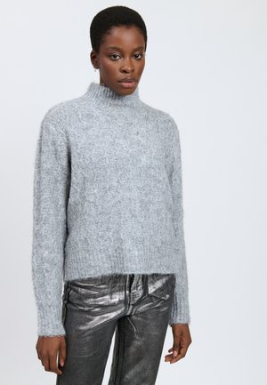 Mujer vistiendo un suéter de cuello alto de punto trenzado en azul claro y pantalones metálicos gris oscuro de cintura alta, contra un fondo liso.