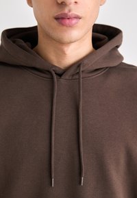 Hnedá mikina s kapucňou a šnúrkou s kovovými koncami. Má mäkkú, hladkú textúru a voľný strih. Detailný pohľad na výstrih a kapucňu.