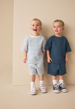 Twee jonge jongens met blond haar staan naast elkaar, een glimlacht en draagt een gestreepte outfit, de ander een effen donkerblauwe outfit.