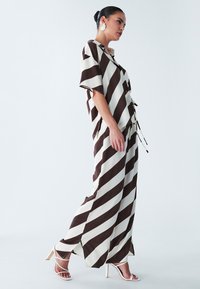Gestreiftes Zweiteiler-Outfit in Braun und Creme, bestehend aus einer kurzärmeligen Bluse und fließenden Hosen mit Seitenschlitzen. Abgerundet durch weiße Absatzsandalen.