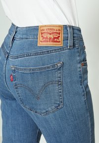Blugi din denim bleu deschis cu talie înaltă, având o pată din piele roșie cu inscripția "Levi Strauss & Co." și cusături distinctive pe buzunarele din spate.
