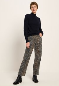 Frau trägt einen schwarzen Rollkragenpullover, Jeans mit Leopardenmuster und schwarzen Knöchelschuhen und steht mit einer Hand in der Tasche.